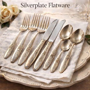 Pode incluir: Conjunto de talheres prateados da Oneida Community, com o design "Enchanted - Gentle Rose". O conjunto inclui garfos, facas e colheres com um padrão floral clássico. A imagem também inclui o texto "Timeless Charm. Vintage Elegance."