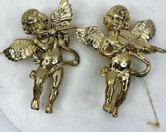 May include: Two gold-coloured cherub figurines. One plays a flute, the other a violin. Both have detailed wings and are in a standing pose. The figurines are likely ornaments or decorative items.