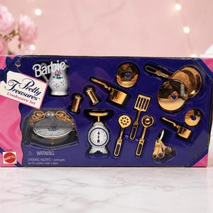 May include: A boxed Barbie Pretty Treasures Cookware Set. The set includes miniature gold-colored pots, pans, and utensils, along with a white and floral-decorated pitcher and a scale. The box is pink and blue.