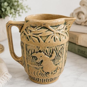 May include: A tan ceramic jug featuring a raised relief design of a duck in a marshy landscape. The jug has a sturdy handle, a pouring spout, and dark green accents within the textured details of the foliage and the duck figure.