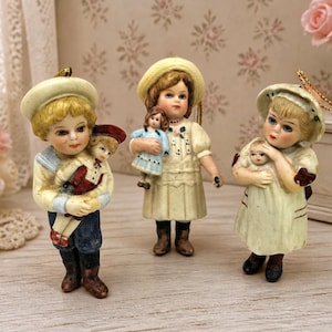 Könnte beinhalten: Drei antike Bisquitpuppen, jede hält eine kleinere Puppe. Die Puppen tragen Hüte und Vintage-Kleidung in Creme, Blau und Rot. Die Puppen stehen auf einer hellen Holzoberfläche mit floralem Hintergrund.