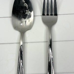 Puede incluir: Un juego de cuchara y tenedor de servir plateados. La cuchara tiene recortes decorativos de estrellas y lunas crecientes. Ambas piezas tienen un diseño floral en los mangos. El juego se muestra sobre una superficie blanca.