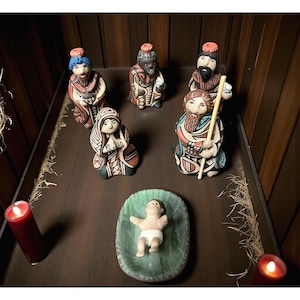 May include: A handcrafted nativity scene with six ceramic figurines and a baby in a green cradle. The figurines include the three wise men, Mary, Joseph, and baby Jesus. The figurines are painted with red, black, and blue accents.