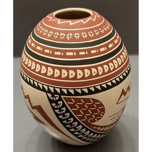 May include: A hand-painted, round ceramic vase with a small opening. The vase features a cream-colored base with intricate designs in red, black, and white. The patterns include geometric shapes and bands, showcasing a traditional aesthetic.