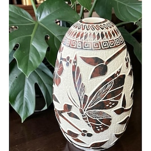 May include: An egg-shaped, off-white vase with a carved design featuring a butterfly, flowers, and leaves in shades of brown. The top rim has a geometric pattern. The vase is a decorative item, likely for display or holding flowers.