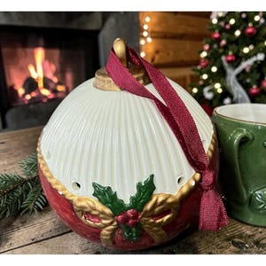 May include: A decorative Christmas ornament with a white, ribbed top and a red base. The base features gold accents, holly, and a bow. A burgundy ribbon is attached for hanging. A green mug is visible in the background.