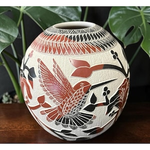 May include: A spherical ceramic vase featuring a textured off-white surface with intricate hand-carved details. The design displays a hummingbird in flight, surrounded by floral motifs and geometric patterns in shades of terracotta red and black.