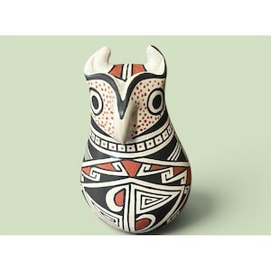 May include: A ceramic owl figurine featuring a teardrop-shaped body decorated with intricate geometric patterns in black, white, and terracotta red. The owl has large circular eyes, a pointed beak, and ear tufts, with detailed dot and line work.