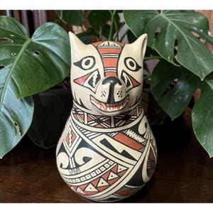 May include: A ceramic wolf figurine with a cream-colored base, adorned with geometric patterns in black and red. The wolf has a smiling expression and pointed ears. The figurine is displayed on a wooden surface with green plants in the background.
