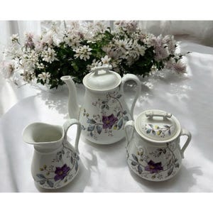 May include: A white ceramic tea set featuring a teapot, creamer, and sugar bowl. Each piece is adorned with a floral pattern of purple flowers and green leaves. The set is designed for serving tea.
