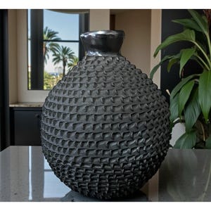 May include: A dark gray, textured vase with a bulbous body and a narrow neck. The surface is covered in a repeating pattern of small, raised shapes. The vase is a decorative home accent.