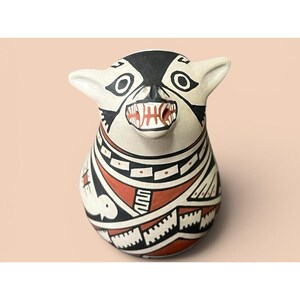 May include: A ceramic vessel shaped like an animal head with pointed ears, featuring a cream-colored base painted with black and terracotta geometric patterns, including a bird motif and a bared-teeth mouth with visible fangs on the front.