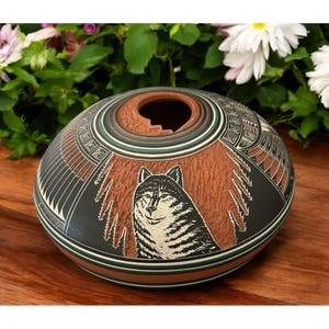 May include: A round, handcrafted ceramic pot with a wolf design. The pot features a brown center with a black and white wolf illustration, surrounded by black and white geometric patterns. The rim has a unique opening.