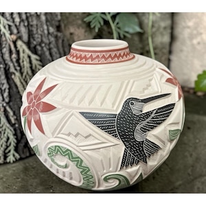 May include: A round ceramic vase with a matte cream finish, featuring hand-carved geometric patterns and relief designs. The surface is decorated with a black hummingbird, red flowers, and green accents, with a patterned band around the neck.