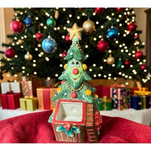 May include: A ceramic Christmas tree figurine with a smiling face, a red nose, and a gold star topper. The tree is decorated with gold beads and colorful ornaments, and it sits atop a square, gift-box-shaped base with a hollow center.