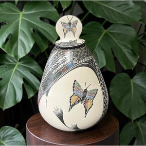 May include: A teardrop-shaped ceramic vessel with a matching lid, featuring a cream-colored base decorated with intricate black geometric patterns and hand-painted butterflies in shades of red, blue, yellow, and green on the body and lid.