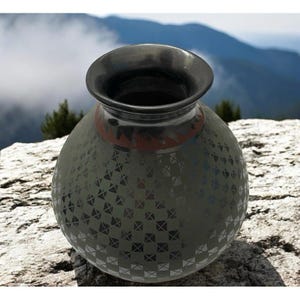 May include: A handcrafted, dark gray ceramic vase with a wide, black rim and a patterned surface. The vase features a geometric design and a band of red and black accents near the top. The vase is displayed outdoors on a rock.