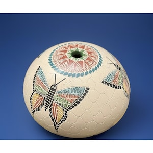 May include: A cream-colored, spherical decorative object with a textured surface. It features a central circular design and two butterflies with colorful wings. The object has a green opening at the top and is set against a blue background.