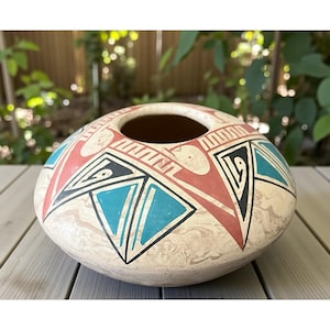 May include: A round, off-white ceramic pot with a wide opening. The pot is decorated with geometric designs in red, turquoise, black, and white. The design includes triangles and other shapes. The pot is sitting on a wooden surface.