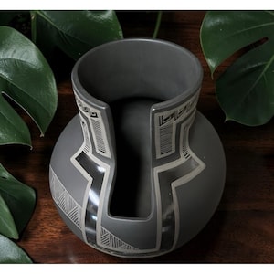 May include: A matte charcoal gray ceramic vessel featuring a wide, circular opening with a deep, U-shaped cutout extending down the neck. The exterior is decorated with intricate, light gray geometric line patterns and angular motifs.
