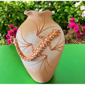 May include: A beige ceramic vase with a ruffled top and a lizard design. The lizard is brown with white spots and is placed on the front of the vase. The vase has brown leaf-like designs and white accents.