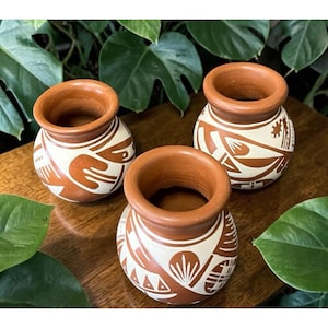 May include: Three small, round terracotta pots with a cream-colored geometric design. The pots have a wide opening and are arranged on a wooden surface, surrounded by green foliage. The pots are likely for decorative purposes.