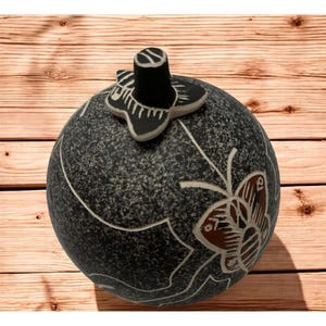 May include: A round, dark gray decorative container with a textured surface and a butterfly design carved into the side. The lid features a black stem and a butterfly-shaped handle. The container sits on a wooden surface.