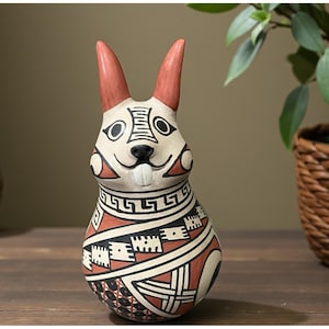 May include: A ceramic rabbit figurine with a hand-painted design. The rabbit has red ears, a black nose, and a white face with black eyes. The body is cream-colored with geometric patterns in red and black. The figurine is on a wooden surface.