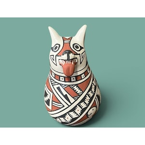 May include: A ceramic dog figurine featuring a teardrop-shaped body painted with intricate geometric patterns in terracotta, black, and white. The dog has pointed ears, a black nose, and an open mouth with a protruding tongue painted in terracotta.