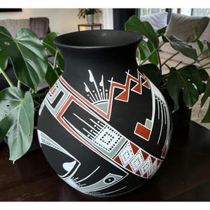 May include: A black ceramic vase with a wide mouth and rounded body. The vase is decorated with geometric patterns in white, red, and gray. The design includes zigzags, squares, and lines, creating a striking visual contrast. The vase is a decorative item.