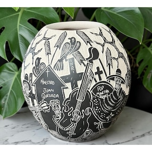 May include: A spherical ceramic vase with a textured cream-colored surface featuring black sgraffito illustrations of skeletons, crosses, and musical instruments. Text includes "Maestro Juan Quezada" and "RIP Martha Ponce" among the detailed carvings.