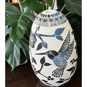 May include: A cream-colored, egg-shaped vase with a detailed hummingbird design. The vase features black and blue accents, with floral and geometric patterns. The hummingbird is depicted in mid-flight, adding a dynamic element to the handcrafted ceramic vase.