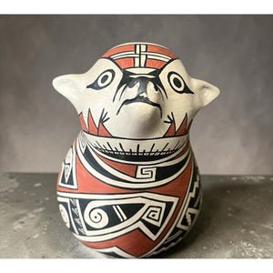May include: A ceramic figurine with a white base, adorned with black and red geometric patterns. The figurine has a stylized animal-like face with large eyes and ears. The design incorporates tribal-inspired motifs.