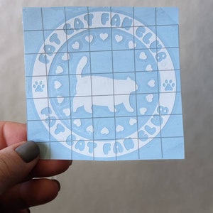 May include: A light blue square sticker with the text "CAT FAN CLUB" in a circle around a white cat silhouette. The design includes hearts and paw prints. The sticker is held by a hand.
