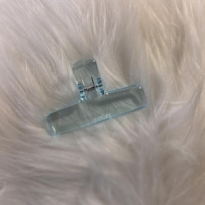 May include: A clear, light blue hair clip with a spring mechanism. The clip is made of transparent plastic and has a rectangular shape with a curved top. The clip is designed to hold hair in place.