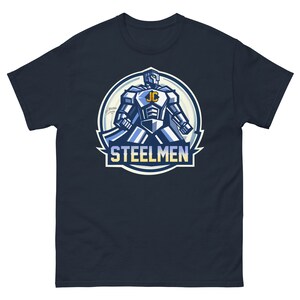 Puede incluir: Camiseta azul marino con un gráfico de un robot azul y plateado que lleva una capa y sostiene un escudo. El robot tiene las letras "JC" en su pecho. El gráfico está rodeado por un círculo azul y blanco con el texto "STEELMEN" debajo.