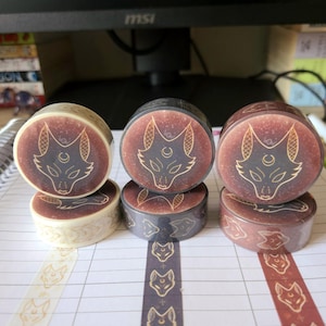 May include: Three rolls of washi tape with a gold wolf design on a brown, black, and white background. The tape is unrolled on a lined notebook page.