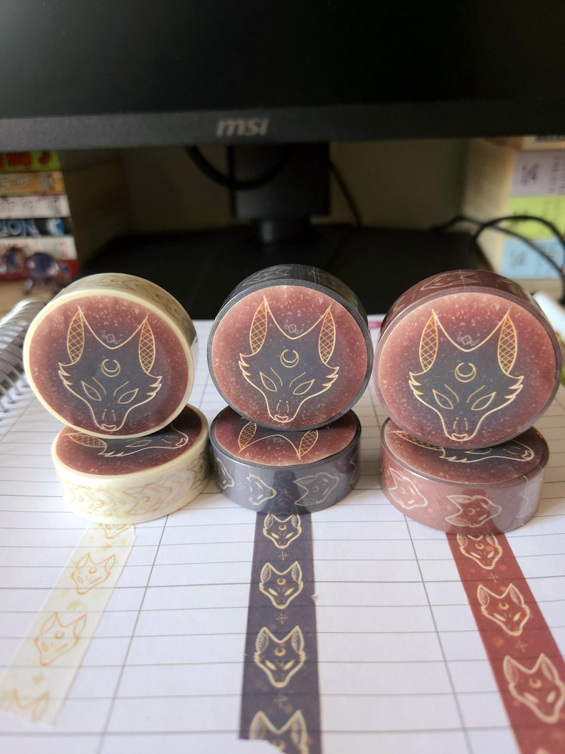 May include: Three rolls of washi tape with a gold wolf design on a brown, black, and white background. The tape is unrolled on a lined notebook page.