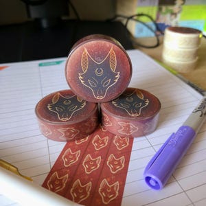 May include: Three rolls of washi tape featuring a repeating pattern of a stylised fox head with a crescent moon and stars. The fox is outlined in gold and the background is a deep red with gold flecks.