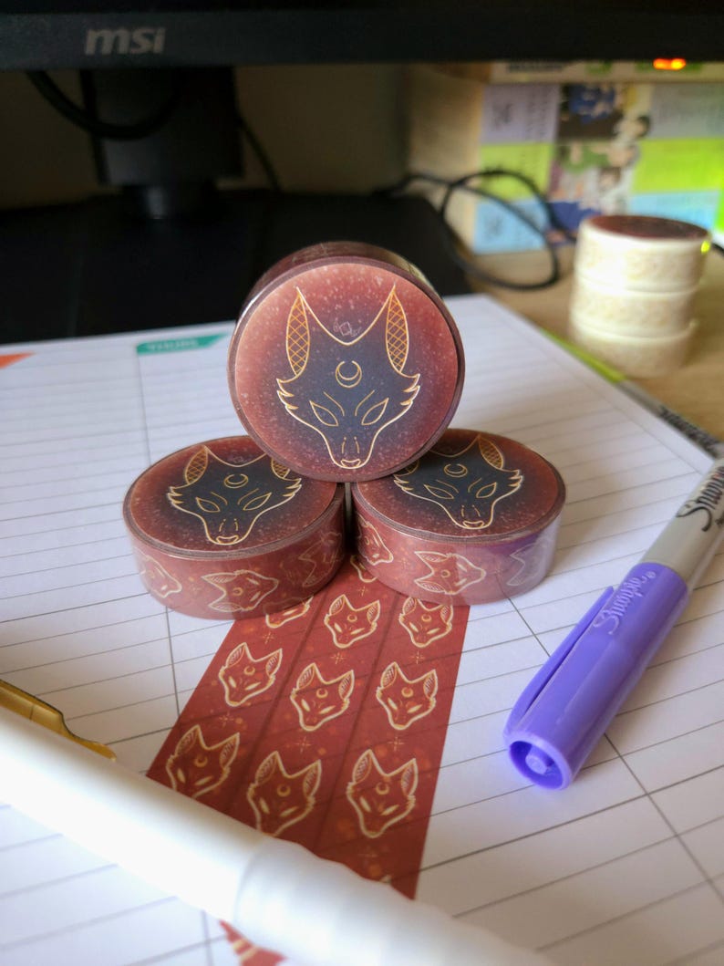May include: Three rolls of washi tape featuring a repeating pattern of a stylised fox head with a crescent moon and stars. The fox is outlined in gold and the background is a deep red with gold flecks.