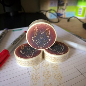 May include: Three rolls of washi tape featuring a gold and black fox design. The tape is on a white lined notebook. The tape is clear and has a repeating pattern of the fox design.