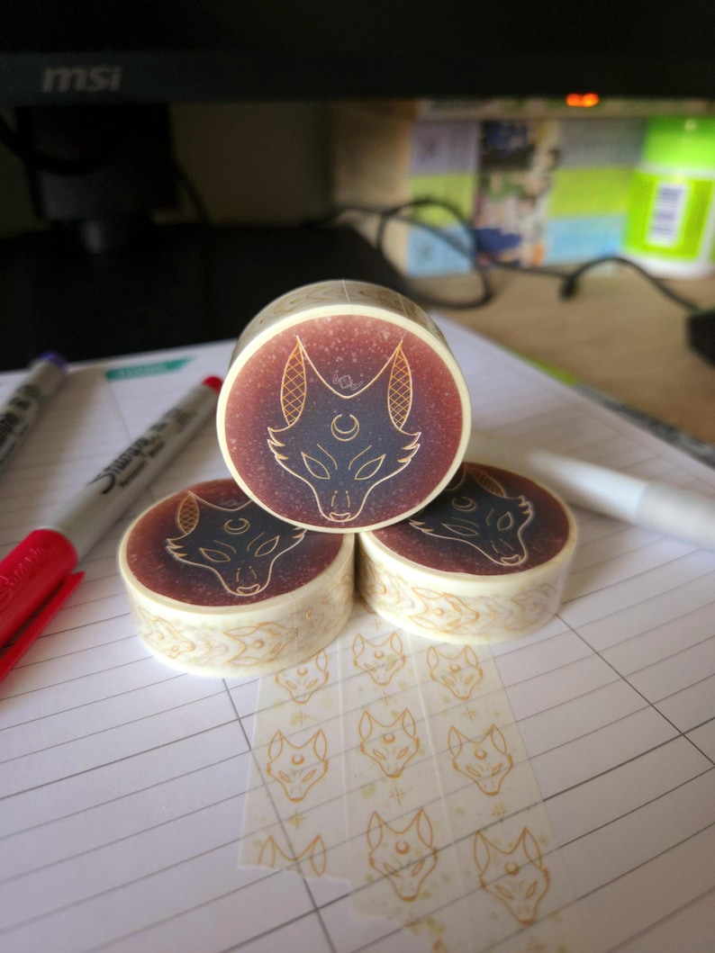 May include: Three rolls of washi tape featuring a gold and black fox design. The tape is on a white lined notebook. The tape is clear and has a repeating pattern of the fox design.