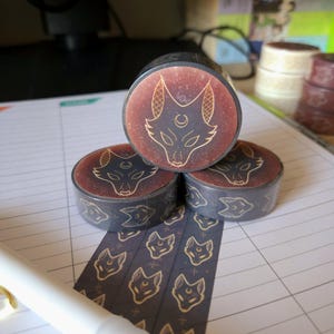 May include: Three rolls of washi tape with a black background and gold foil design of a wolf's head with a crescent moon. The tape is unrolled and shows a repeating pattern of the wolf's head.