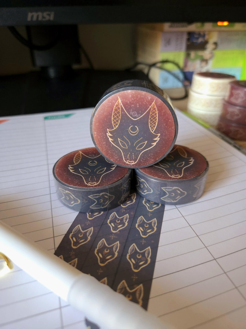 May include: Three rolls of washi tape with a black background and gold foil design of a wolf's head with a crescent moon. The tape is unrolled and shows a repeating pattern of the wolf's head.