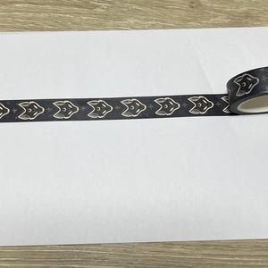 May include: Black and gold washi tape with a repeating pattern of fox faces. The tape is rolled up and partially unrolled on a white surface.