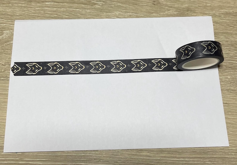 May include: Black and gold washi tape with a repeating pattern of fox faces. The tape is rolled up and partially unrolled on a white surface.