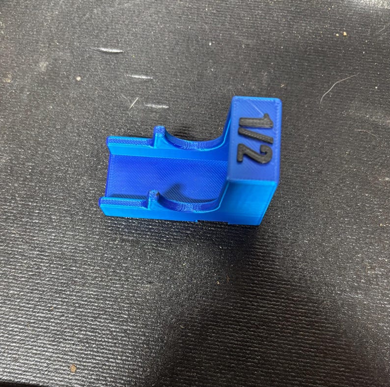 Puede incluir: Un objeto de pl&aacute;stico azul brillante, impreso en 3D, con el n&uacute;mero "1/2" impreso en negro. El objeto tiene forma rectangular con un interior curvo y dos peque&ntilde;as protuberancias. Probablemente sea una herramienta o un componente.