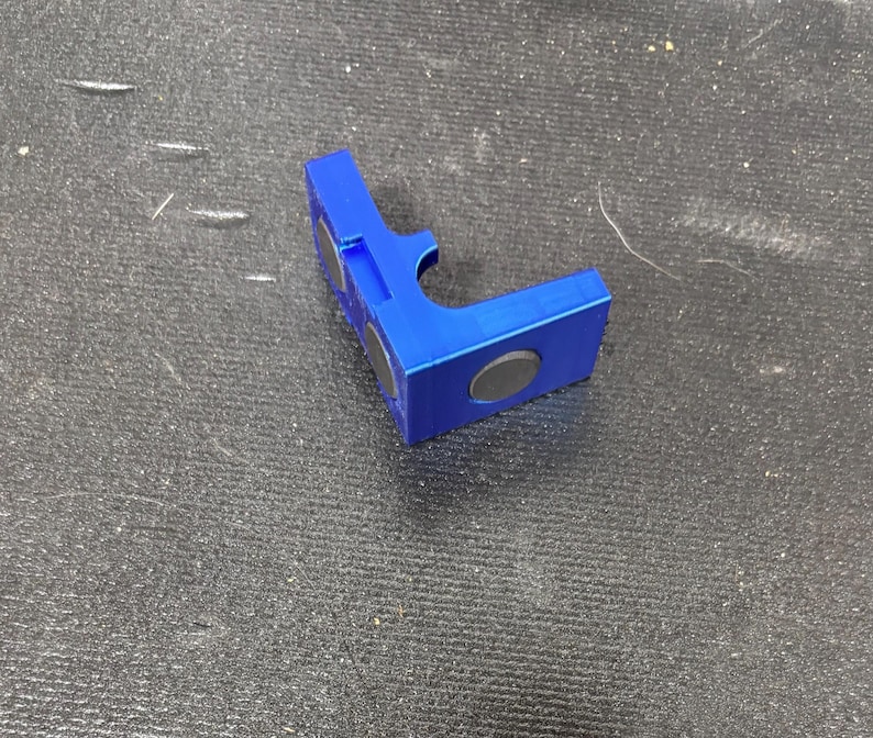 Puede incluir: Un objeto azul brillante en forma de L con dos imanes negros circulares adheridos. El objeto tiene un recorte curvo en un lado y parece estar hecho de pl&aacute;stico o un material similar. Descansa sobre una superficie oscura y texturizada.