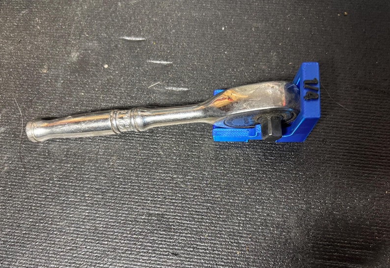 Puede incluir: Una llave de carraca plateada con un soporte azul de 1/4 de pulgada. La llave est&aacute; sobre una superficie negra texturizada. El soporte azul tiene el n&uacute;mero "1/4" impreso.