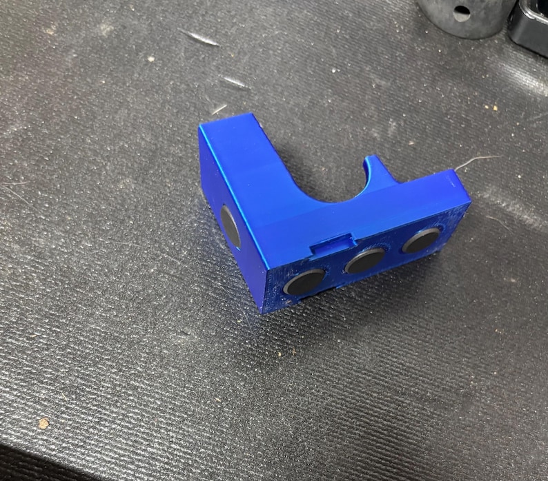 Puede incluir: Un soporte azul brillante en &aacute;ngulo recto con tres imanes negros integrados. El soporte tiene una secci&oacute;n curva y una ranura. El objeto est&aacute; hecho de un material s&oacute;lido y se encuentra sobre una superficie oscura texturizada.